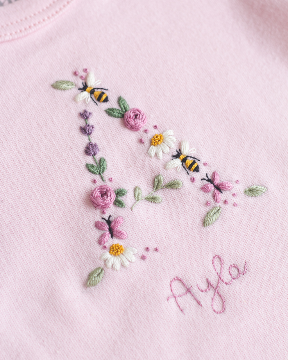 Tecido rosa com o nome Ayla, flores e abelhas bordadas.