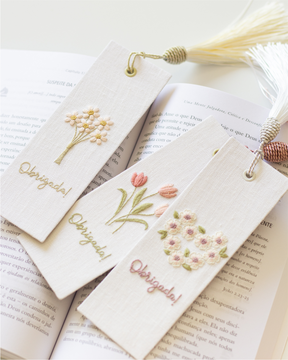 Três marcadores de tecido bordados com flores e a palavra Obrigada, sobre um livro aberto.